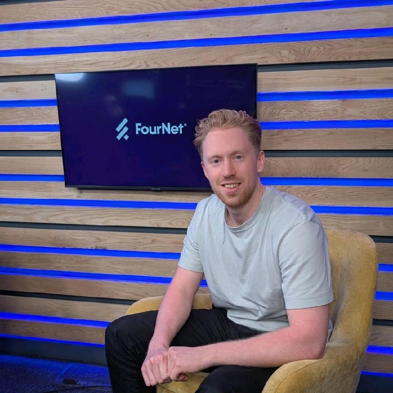 Bill Cooper, a white man sat on a chair in front of a screen displaying the FourNet logo.